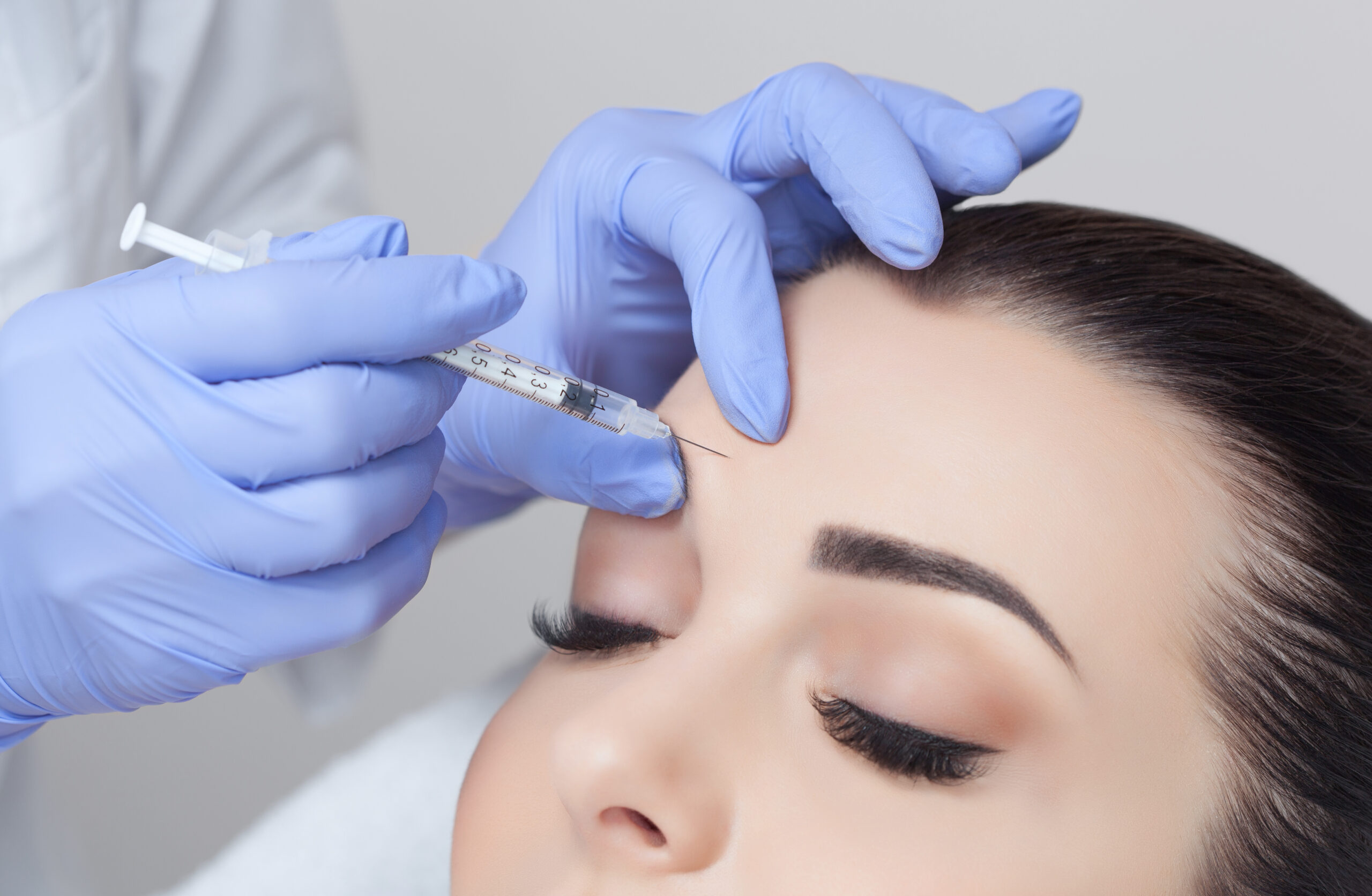 Close-up of a cosmetic procedure where a healthcare professional in blue gloves administers an injection to the forehead of a woman receiving a beauty treatment, highlighting the use of a syringe for aesthetic enhancements.