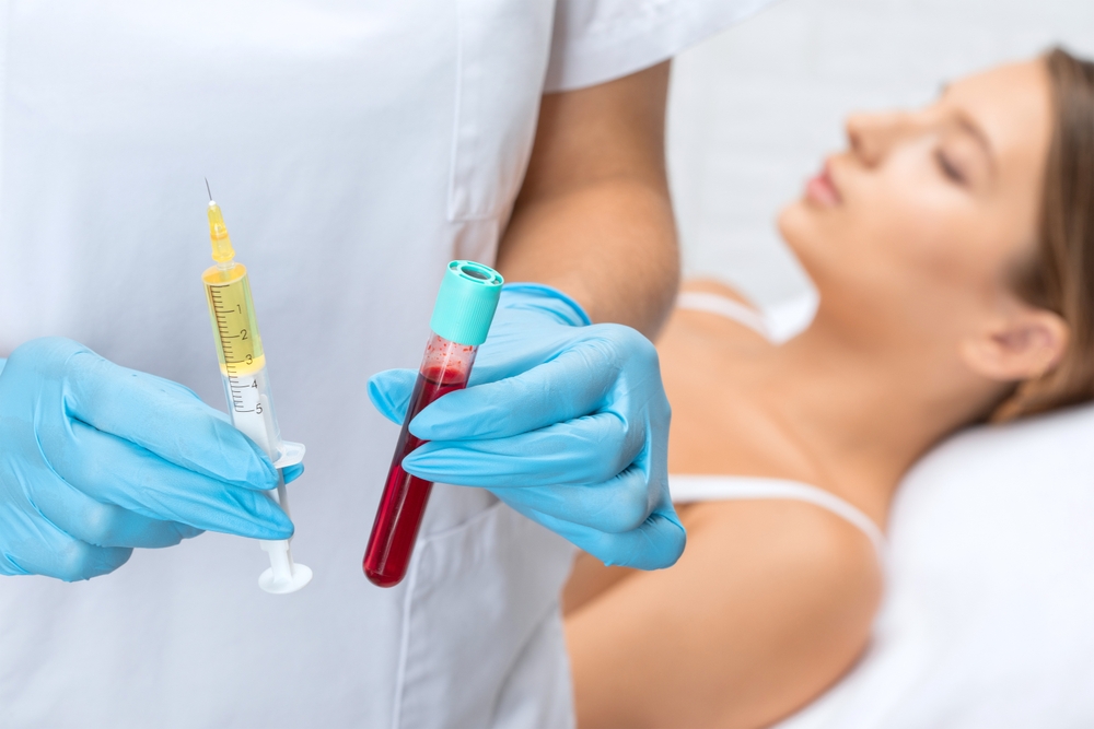 Healthcare professional in blue gloves holding a syringe filled with yellow liquid and a blood collection tube while a patient rests in the background, illustrating a medical procedure.