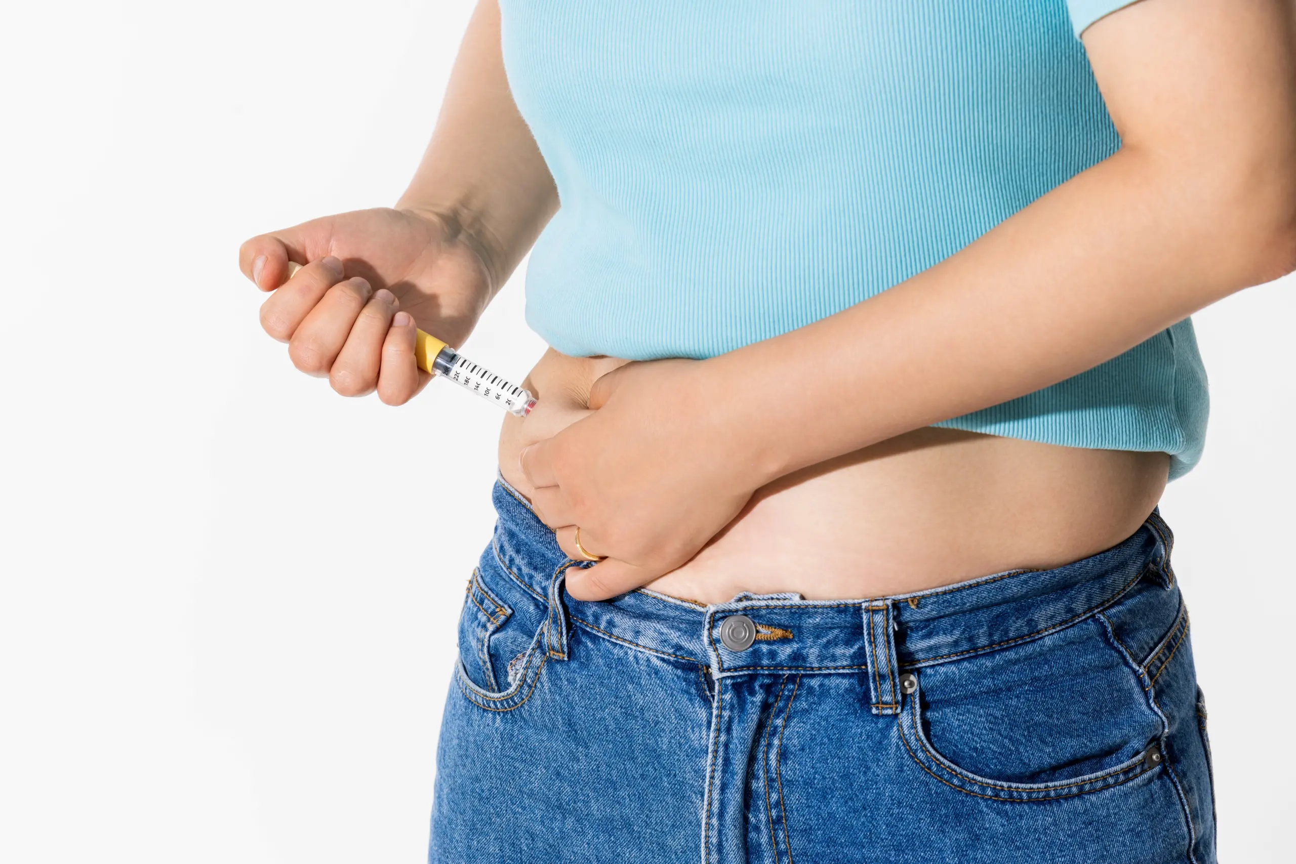 A person administering an injection into their abdomen, wearing a light blue top and denim jeans, demonstrating self-injection techniques for diabetes management or medication administration.
