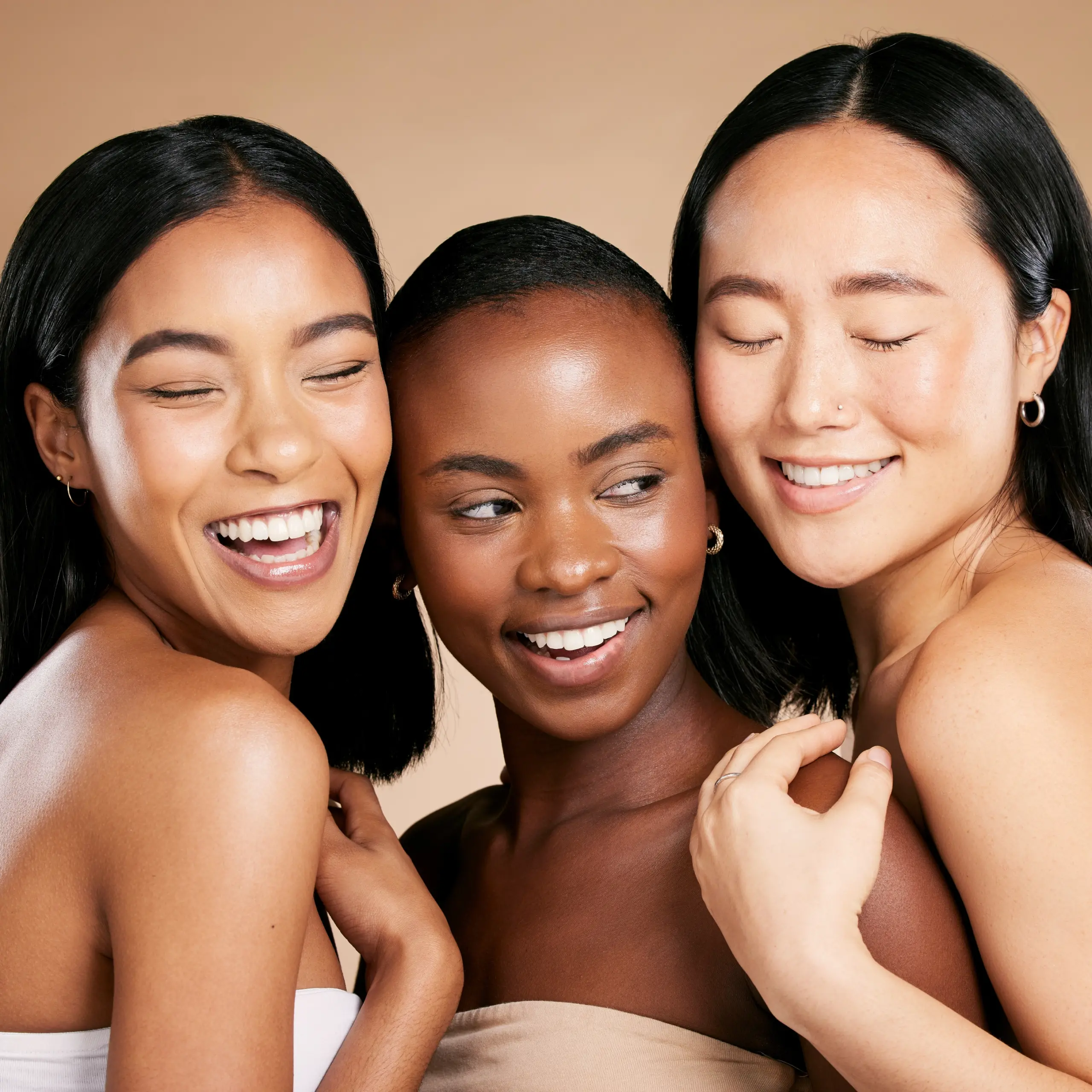 Three diverse women with radiant smiles pose closely together against a soft beige background, showcasing natural beauty and friendship.