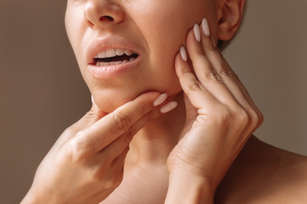 Close-up of a woman touching her jawline with both hands, indicating discomfort or pain, showcasing her expression of concern. The background is neutral, emphasizing her facial features and the action of self-examination.