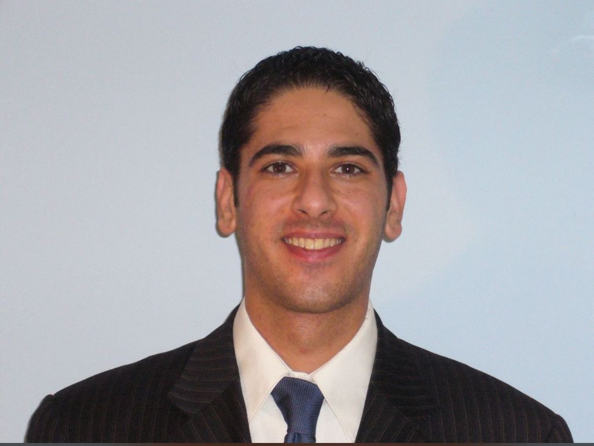 Professional headshot of a smiling young man in a formal suit and tie, set against a light blue background, suitable for business profiles or professional networking.