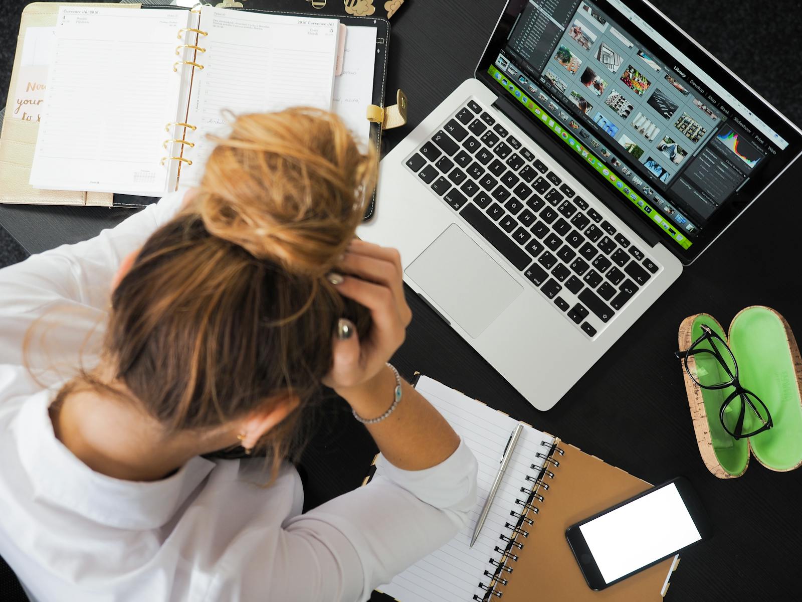 A woman sitting at a desk with a laptop, planner, and notebook, appearing stressed while holding her head. Nearby, a smartphone and glasses are visible, indicating a busy work environment.