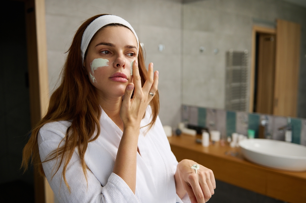 Woman applying a green facial mask in a modern bathroom, wearing a white bathrobe and headband, with skincare products visible in the background.