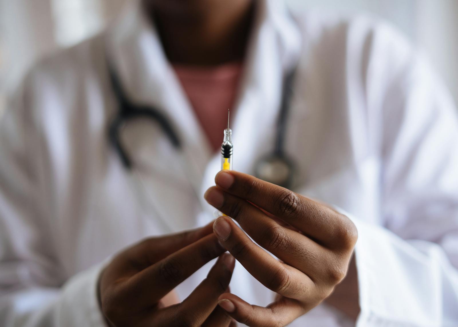Healthcare professional holding a syringe with a yellow liquid, wearing a lab coat and stethoscope, symbolizing vaccination or medical procedures.