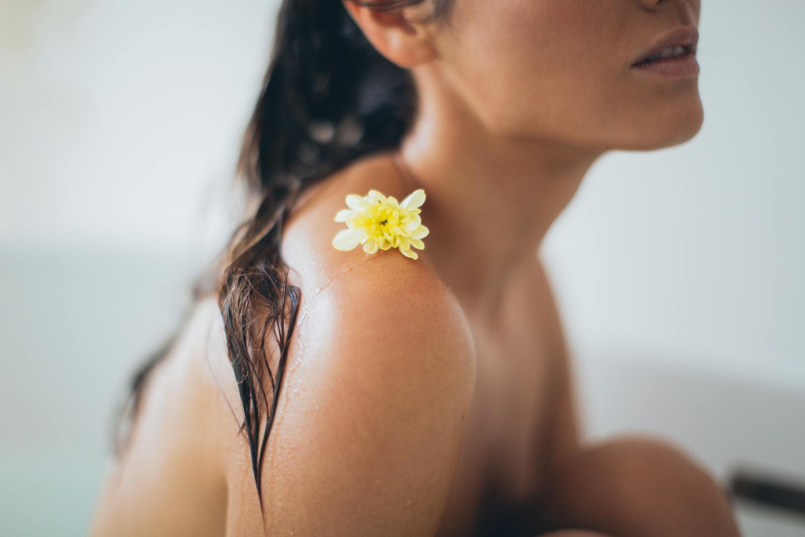 Close-up of a woman with wet hair and a yellow flower on her shoulder, sitting in a serene bath setting, emphasizing relaxation and natural beauty.
