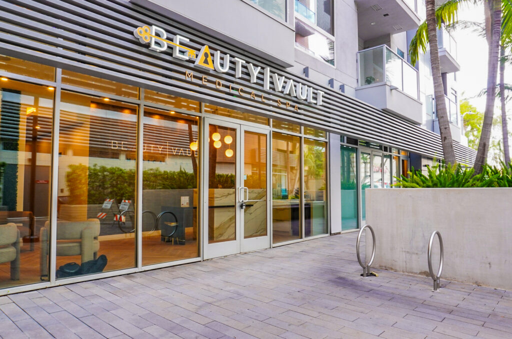Entrance of Beauty Vault Medical Spa featuring large glass doors and modern signage, surrounded by lush greenery and contemporary architecture.