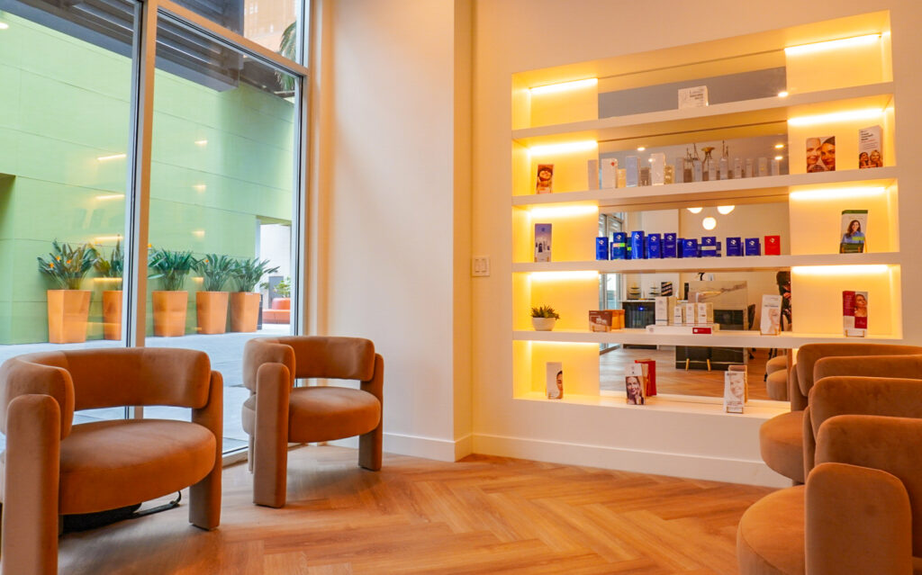 Interior view of a modern skincare clinic featuring two comfortable chairs, a bright display shelf with various skincare products, and large windows revealing a landscaped outdoor area with potted plants.
