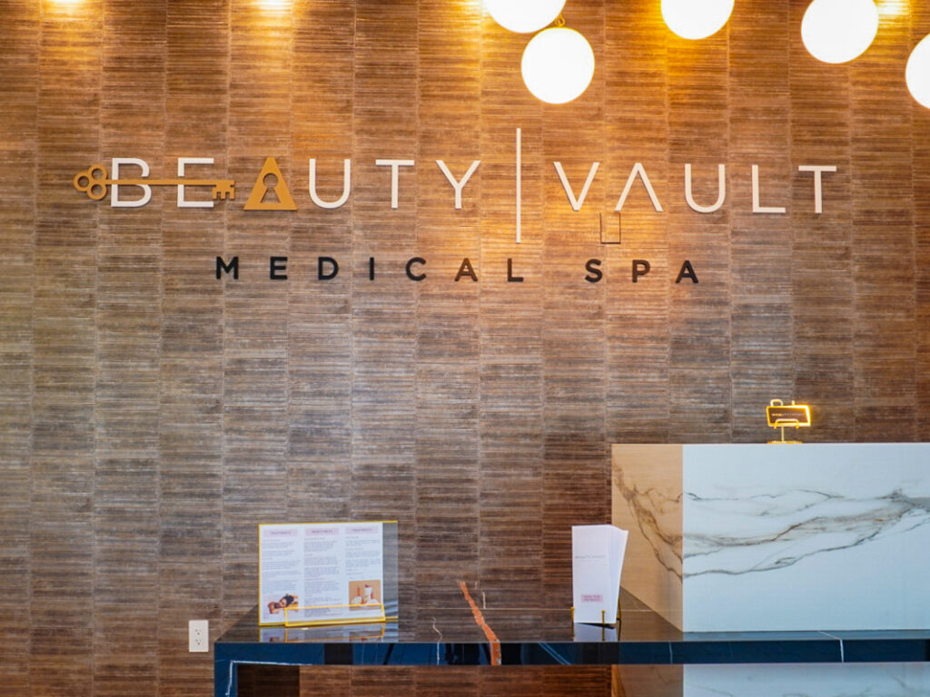 Interior view of Beauty Vault Medical Spa featuring a stylish reception area with modern design elements, including a large illuminated sign and informational brochures displayed on a sleek countertop.