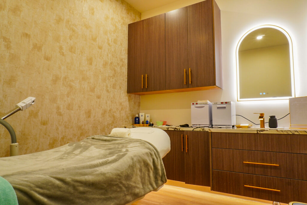 Interior view of a modern treatment room featuring a massage table draped with a soft blanket, wooden cabinetry with gold handles, and a well-lit mirror. The space is designed for relaxation, equipped with beauty products and tools, reflecting a serene atmosphere.