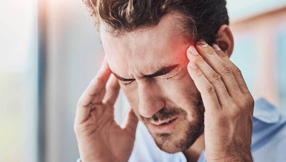 Man suffering from a severe headache, holding his temples with a pained expression, indicating discomfort or migraine symptoms in a bright, indoor setting.